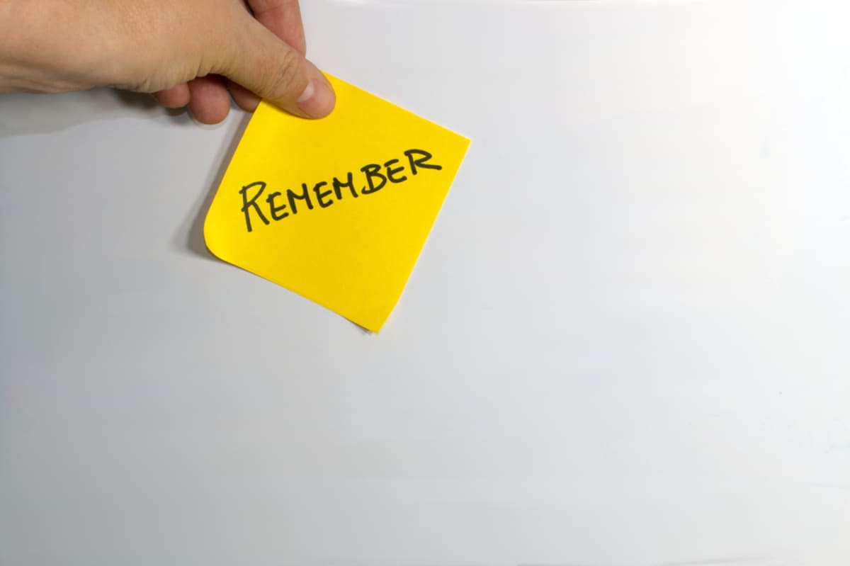 Left hand holding a yellow adhesive note with the word REMEMBER in white background